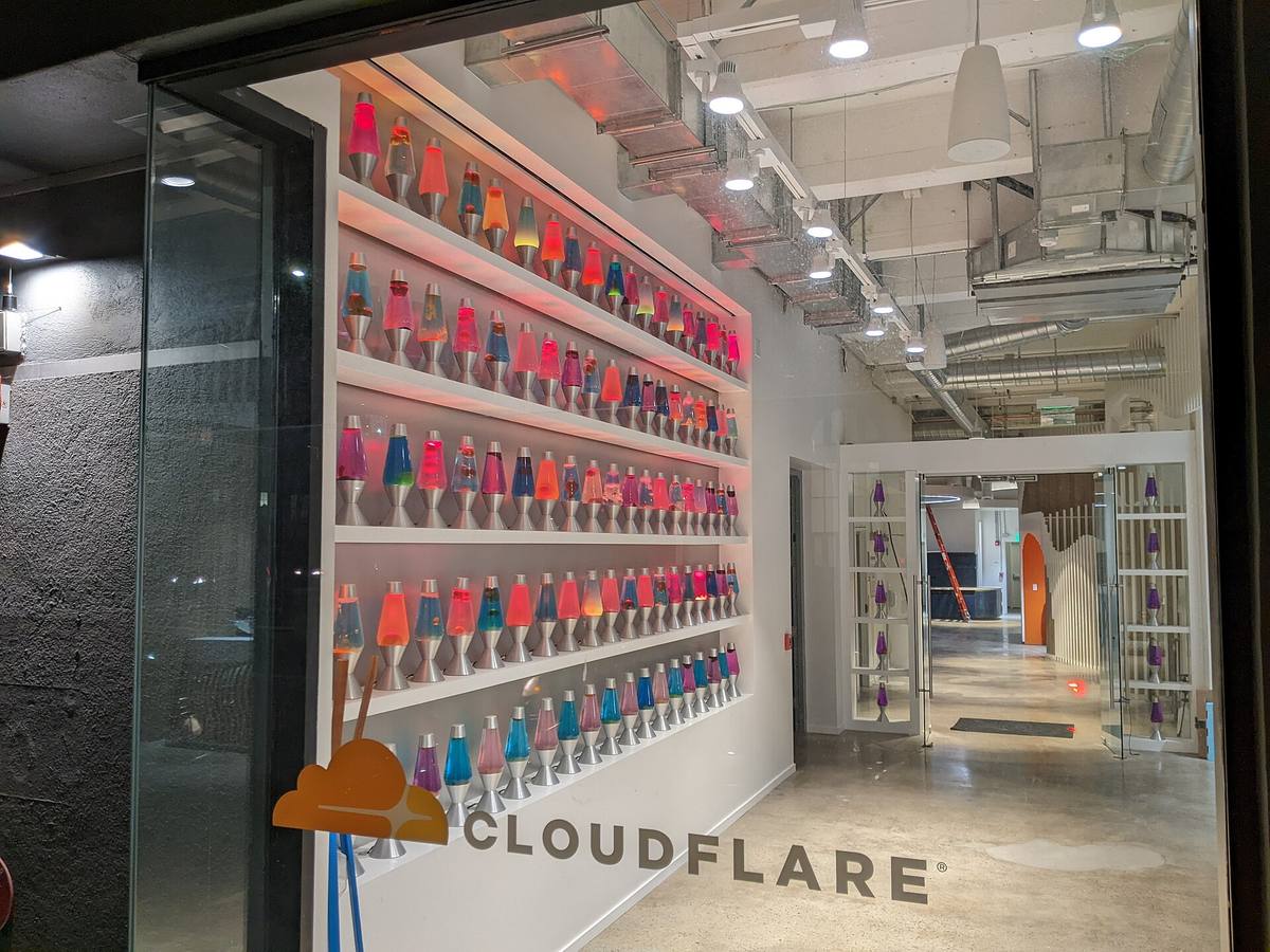Stěna s regály plnými barevných lávových lamp ve vstupní hale, na zdi pod nimi nápis Cloudflare