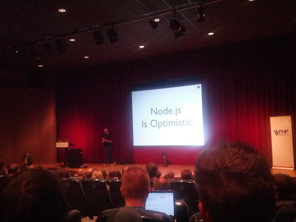 Zatemněný sál s plátnem, na kterém je promítnutý snímek s nápisem Node.js Is Optimistic